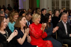 A crowd of people wearing formal clothes seated at an indoor event. They are all looking ahead, smiling and applauding.