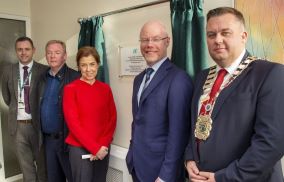 Group of 5 people standing in front of a plaque on a wall. There are green curtains on eithe side of the plaque.