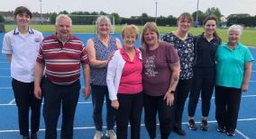 Large group of people standing in a line on an athletics track. There are a couple of healthcare workers with the group. 