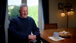 A man is sitting a table in a home setting looking relaxed. His elbow is resting on the table and he is smiling. 