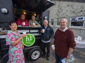 3 people, 2 men and a woman standing before a coffee tricks with coffee cups in their hands. A healthy Ireland logo is on the coffee truck and 2 women are inside the coffee truck.
