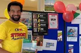 A man wearing a stroke awareness t-shirt, holding a booklet and standing beside a stand covered in stroke awareness graphics.
