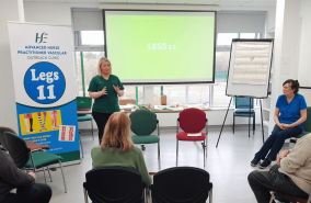 Healthcare perofessional standing giving a talk to a group of peoplein a room. There is a large screen and a pull up stand which reads 'Legs 11' in the background. 