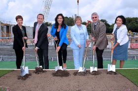 Six people in formal clothes holding shovels over a plot of dirt outdoors.