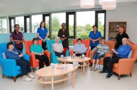Six older people sitting in armchairs indoors, with five healthcare workers standing behind them.