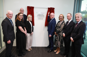 Seven people in formal clothes standing beside Minister for Health Stephen Donnelly, who stands in front of a pair of small red curtains and a sign on a wall 