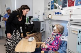 A woman standing and speaking to an older patient in a clinical setting. The patient has a white patch over her eye and there is medical equipment on either side of her. Behind them is a healthcare worker in blue medical clothes.