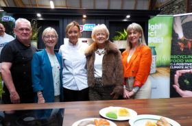 A woman in a chef's uniform stands with four people in regular clothes behind plates of food on a countertop.