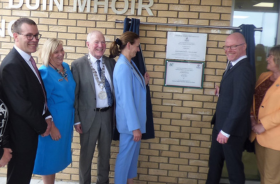 Minister Stephen Donnelly standing beside a plaque on the wall of a healthcare setting with two other men and three women wearing formal clothes.