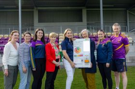 Eight people standing on a football pitch. Two in the centre hold a board promoting skin cancer awareness.