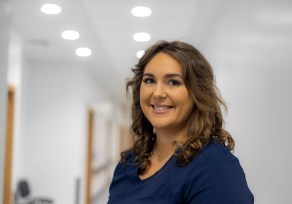 A nurse wearing blue scrubs is standing in the middle of a hospital corridor. She is smiling brightly. 