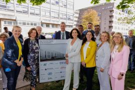 Large group of people standing around a sign depicting an image of a building. There are tall building in the background. 