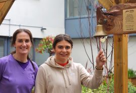 University student Nora Goaley and a nurse standing outside in a pergola area. Nora holds a bell which she is about to ring. 