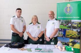 Three National Ambulance Service staff standing behind a table with first aid material on it, beside them is a National Ambulance Service pull-up poster.