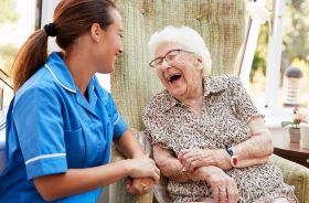 A healthcare worker sitting beside an older woman, both looking at each other and smiling.