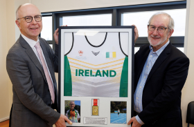 Two men stand holding a framed Ireland sports shirt with a gold medal displayed beneath it.