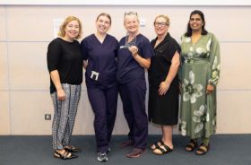Five women standing in a row and smiling. Two in the centre are wearing medical clothes and one is holding a small plaque.