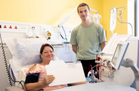 A woman sitting up in a hospital bed in a clinical setting and a man standing beside her.