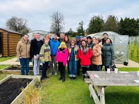 Large group of people standing in a garden setting. There are raised beds in front of the group and 2 polytunnels behind them.