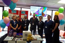 Seven hospital staff members stand behind a table with various informational materials on it. There are baloons beside them and more informational material on a board behind them.