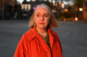 A woman with grey hair and a red coat standing outside with some houses and trees in the background.