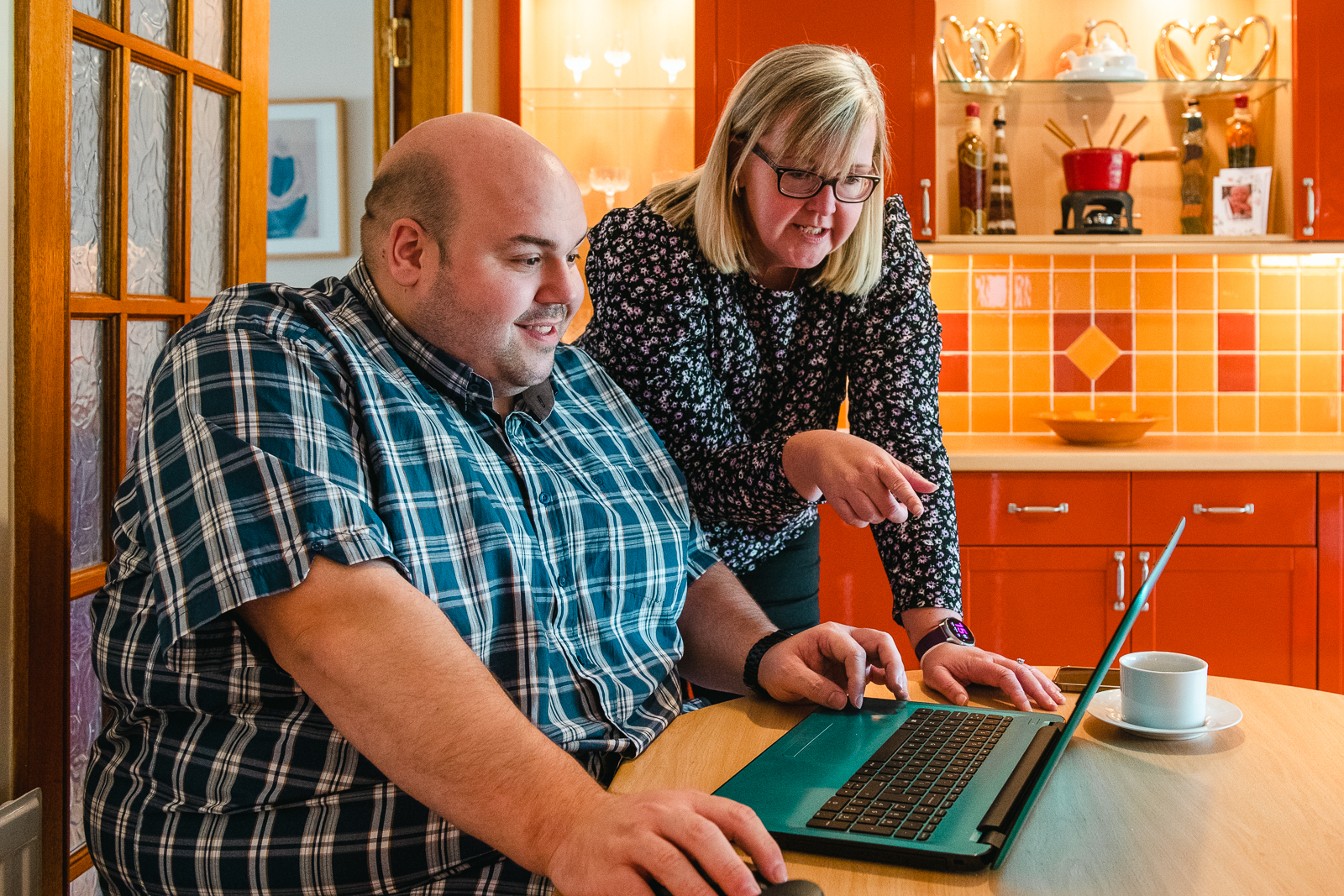 A man sits behind a laptop at a kitchen table. A woman stands beside him also looking at the laptop screen.